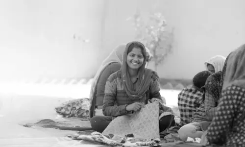 A woman sits on the ground with a joyful smile while stitching fabric, surrounded by other women in the background, all engaged in a workshop. The black-and-white image highlights the positive energy and empowerment of women working together.
